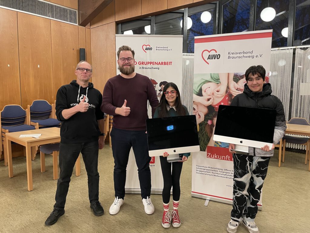 Zwei Kinder, Jörg Elmenthaler und Jonas Wilke stehen lächelnd in einem Raum mit AWO-Bannern im Hintergrund. Die Kinder halten zwei iMacs.